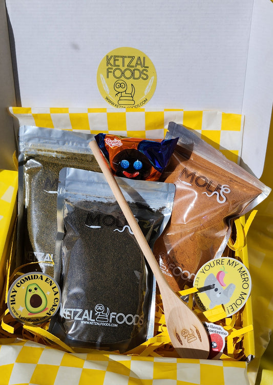 A gift box from Ketzal Foods containing three packets of mole sauce and a wooden spoon with a Ketzal Foods logo on it, all presented on a yellow checkered background.